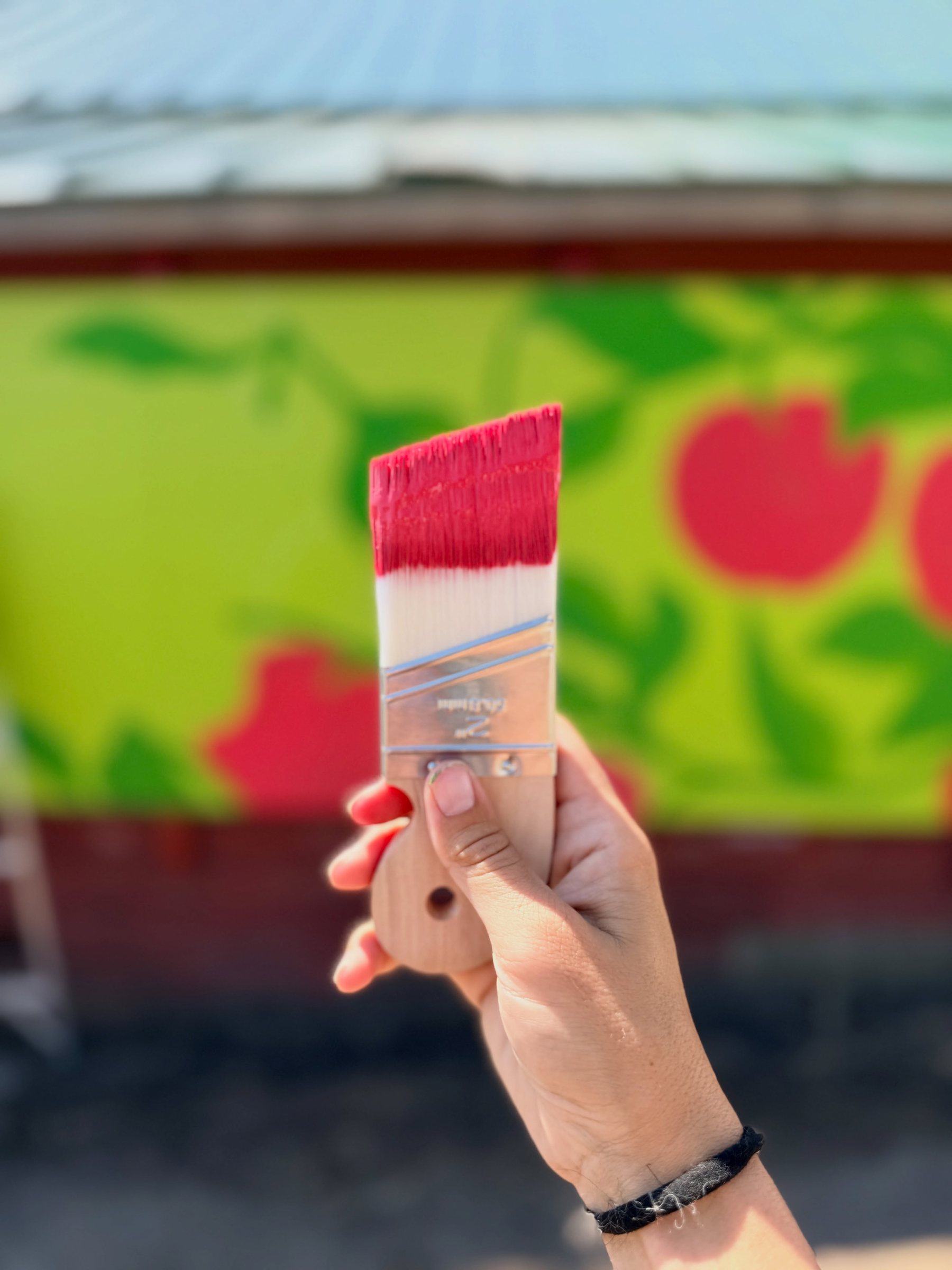 Close-up of a paintbrush loaded with red paint against the mural background